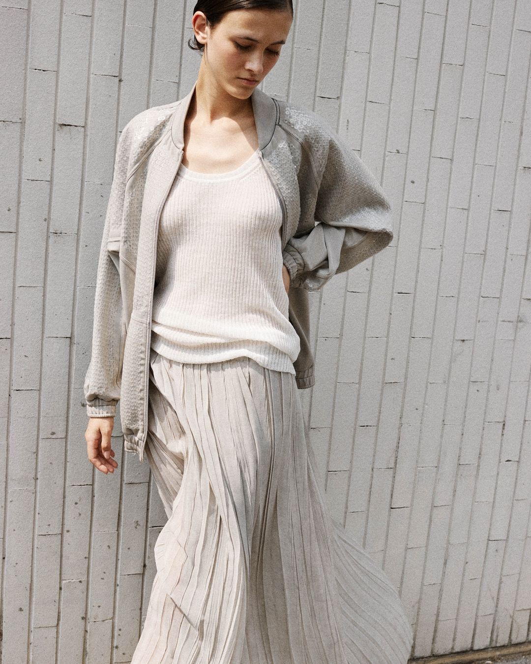 Woman in gray jacket and pleated skirt poses against a textured wall, showcasing a casual and modern fashion ensemble with natural light.