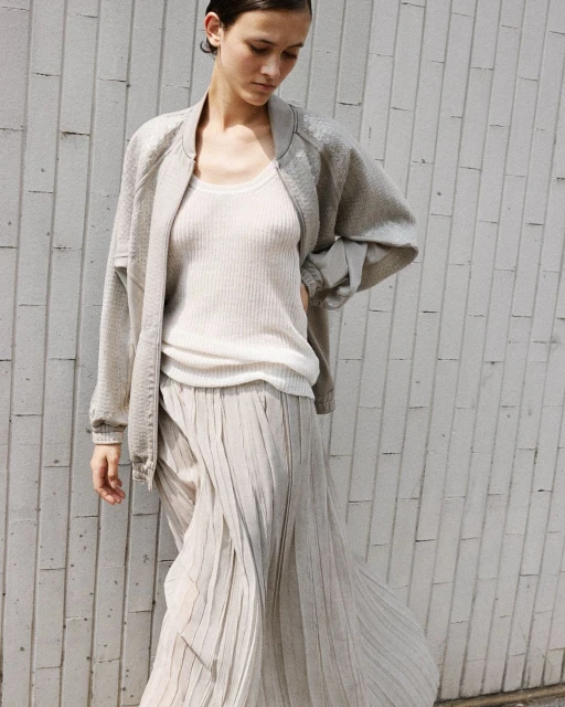 Woman in gray jacket and pleated skirt poses against a textured wall, showcasing a casual and modern fashion ensemble with natural light.