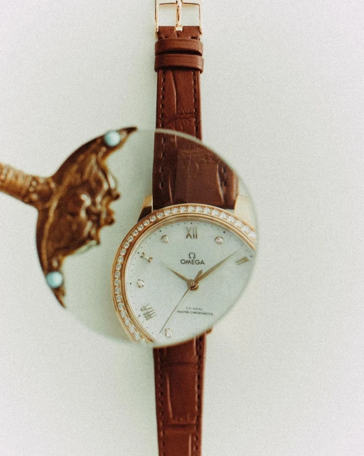 Elegant Omega De Ville watch on a brown leather strap, partially obscured by a reflective surface, set against a clean white background.