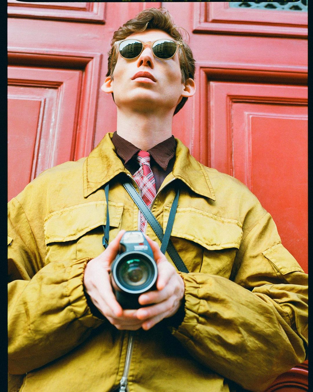 Young adult man with sunglasses holds camera in front of red panel door, wearing a yellow jacket, in a medium shot.