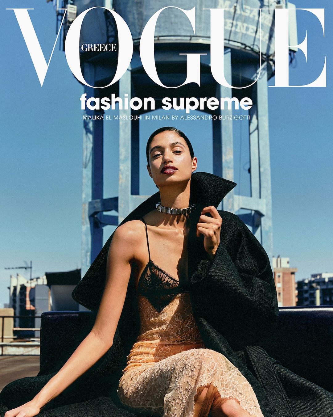 Vogue Greece cover featuring a woman in a slip dress and black coat, seated against a rooftop and water tower backdrop under a blue sky.