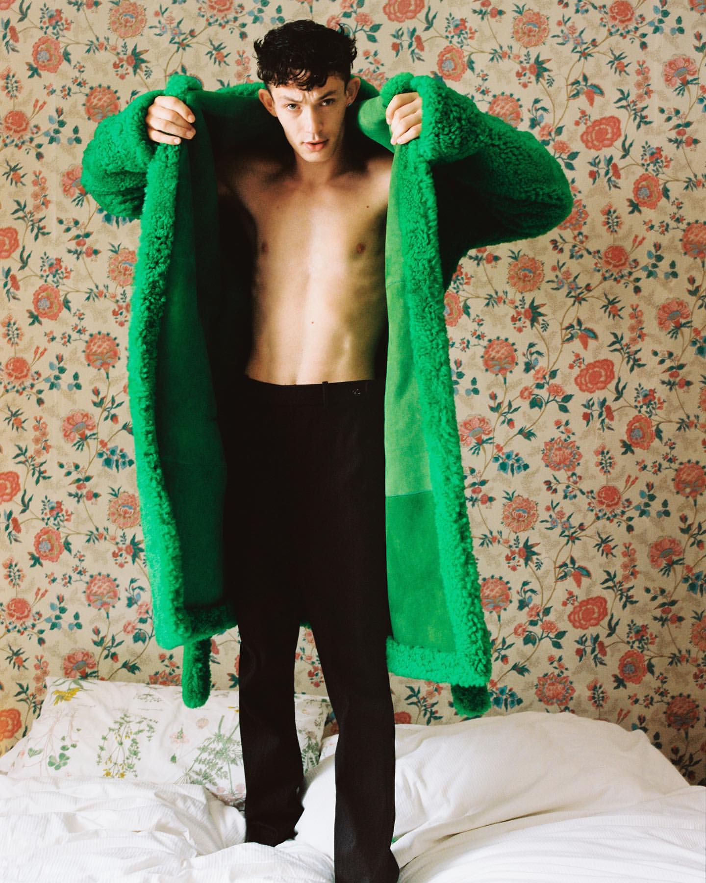 Man with bare chest wearing a green fuzzy coat standing on a bed in a floral patterned room. Fashion editorial shot, indoor.