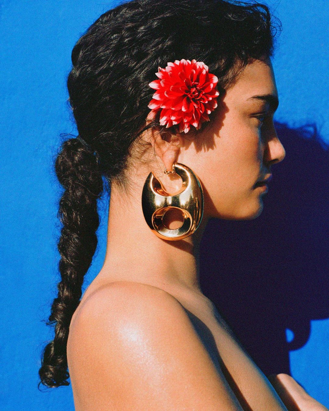 Portrait of a young woman with a braided ponytail and a large gold earring. A red flower adorns her hair against a blue background.