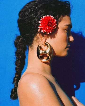 Portrait of a young woman with a braided ponytail and a large gold earring. A red flower adorns her hair against a blue background.