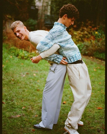 Two young men stand outdoors on a grassy lawn, playfully back-to-back. One smiles, wearing a white sweater, while the other is in a blue knit.