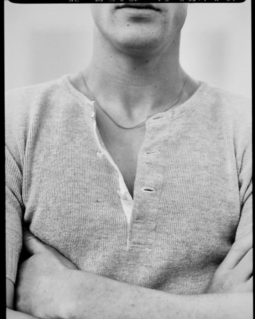 Black and white portrait of a man with folded arms wearing a long-sleeved Henley shirt and a necklace, featuring a neutral background.