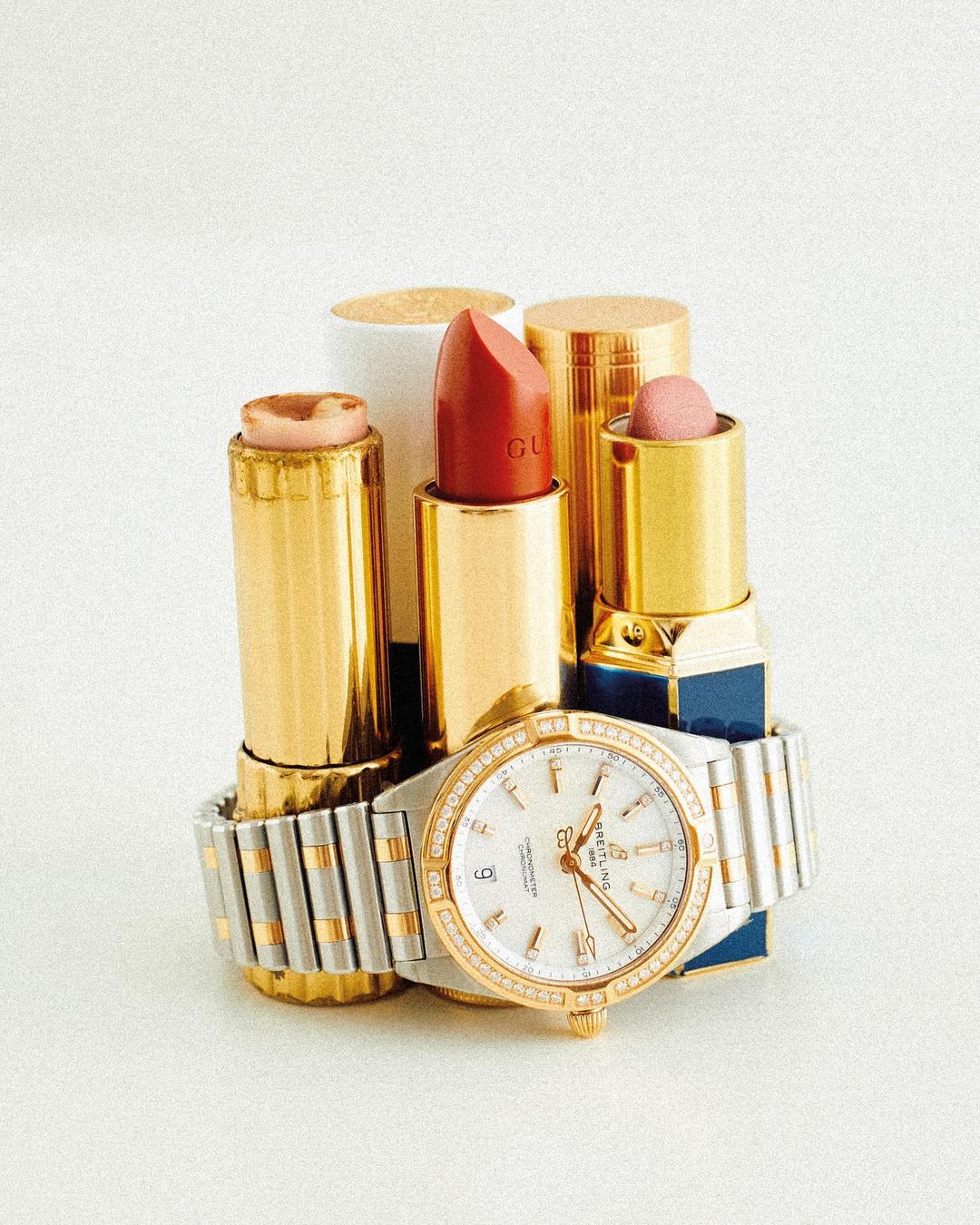 Close-up of gold lipstick tubes stacked with a stainless steel and gold watch on a seamless white background in a studio setting.