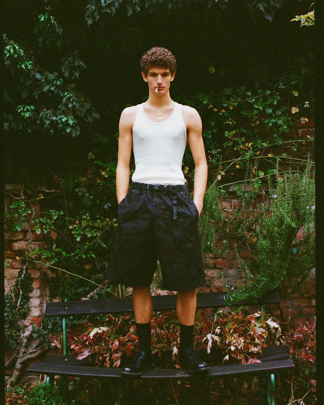Young man in a white tank top and dark shorts standing outdoors in front of lush green foliage and a brick wall.