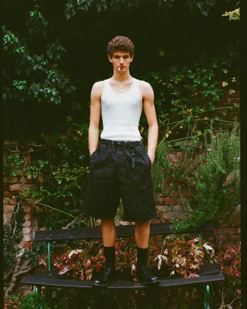 Young man in a white tank top and dark shorts standing outdoors in front of lush green foliage and a brick wall.