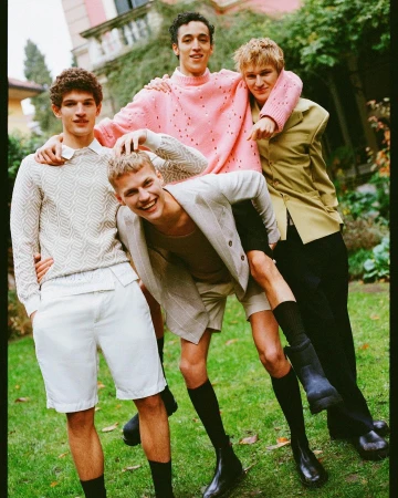 Four stylish young men are having fun outdoors in a group, showcasing contemporary menswear fashion and youthful energy on green grass.
