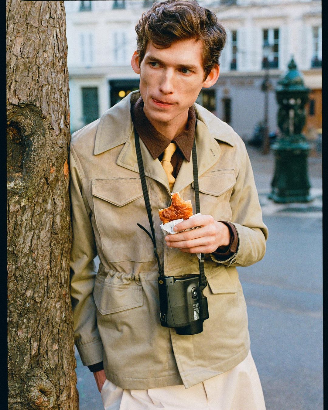 Adult male in a khaki jacket stands next to a tree in an urban environment, holding a croissant, with a camera around his neck.