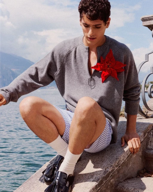 Male model poses seated near water wearing a grey sweater, striped shorts, socks, and loafers with a large red poinsettia pin.