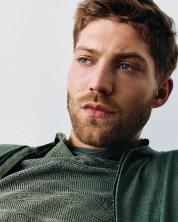 Close-up portrait of a man with short brown hair and a beard wearing a green textured zip-up sweater against a white background.