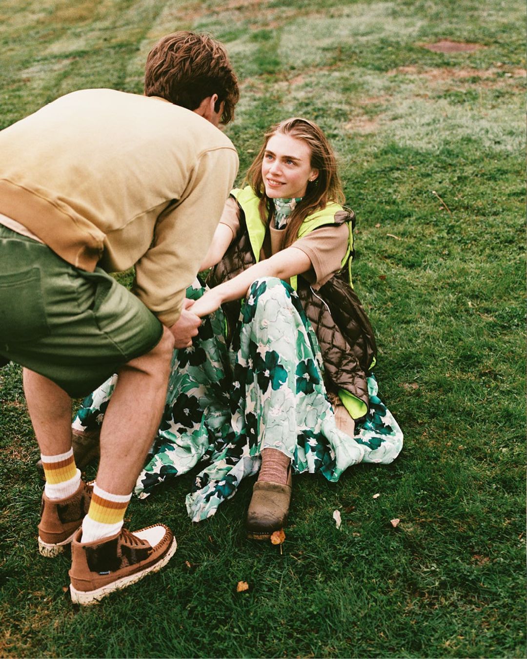 A woman sitting on grass in a floral dress is being helped up by a man in a tan sweatshirt and green shorts in a daytime outdoor setting.