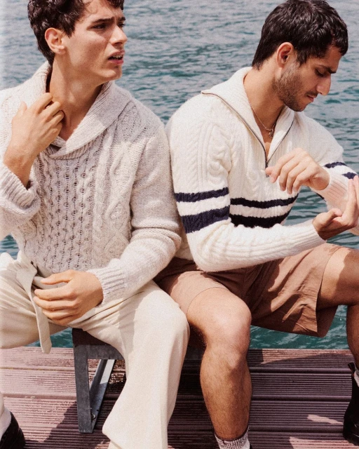 Two adult men sit on a pier wearing sweaters and shorts with a lake visible in the background in a medium shot at eye level.