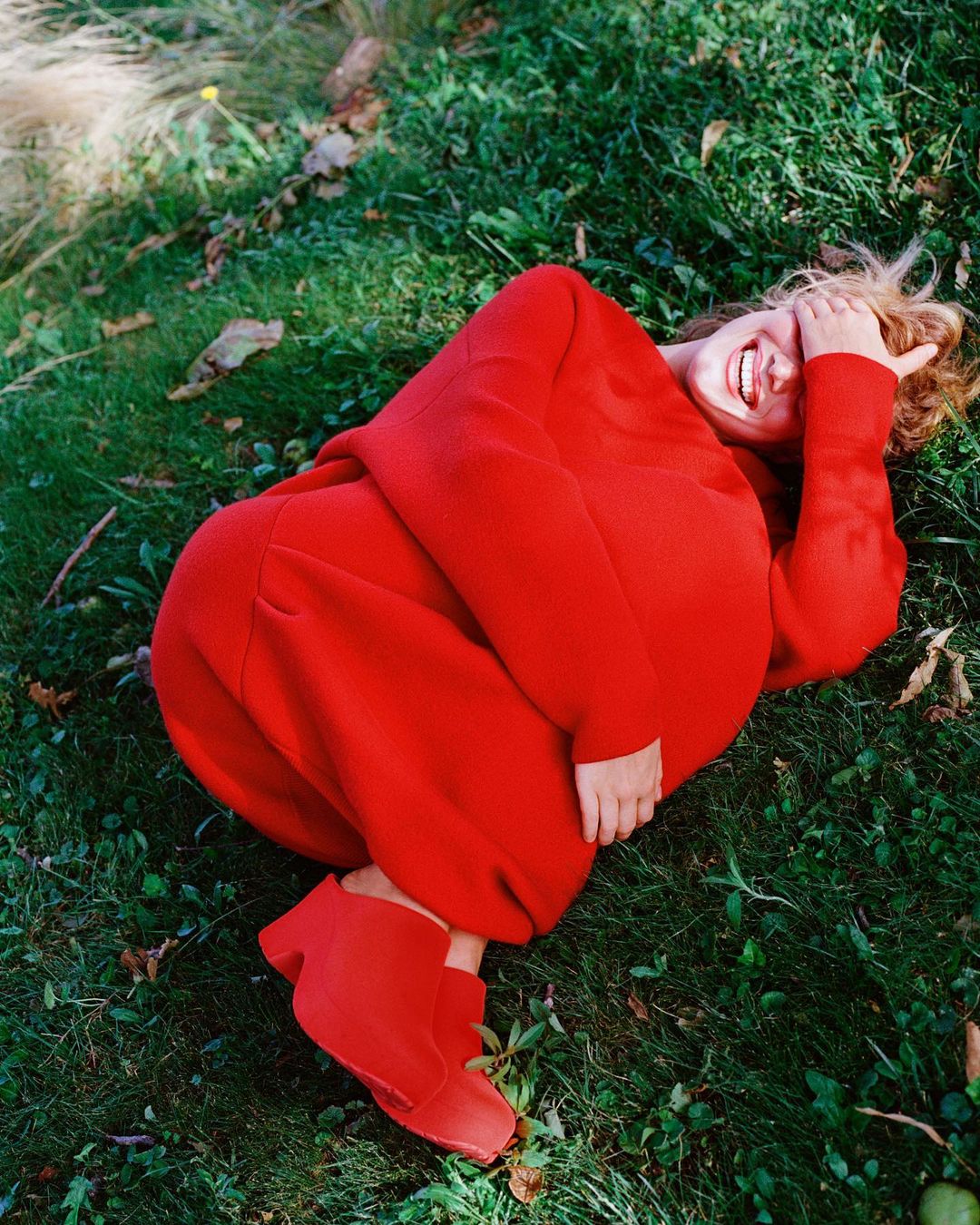 Woman in a red outfit reclines on grass. She smiles while covering her face. Grass and leaves fill the image to the edges of the frame.