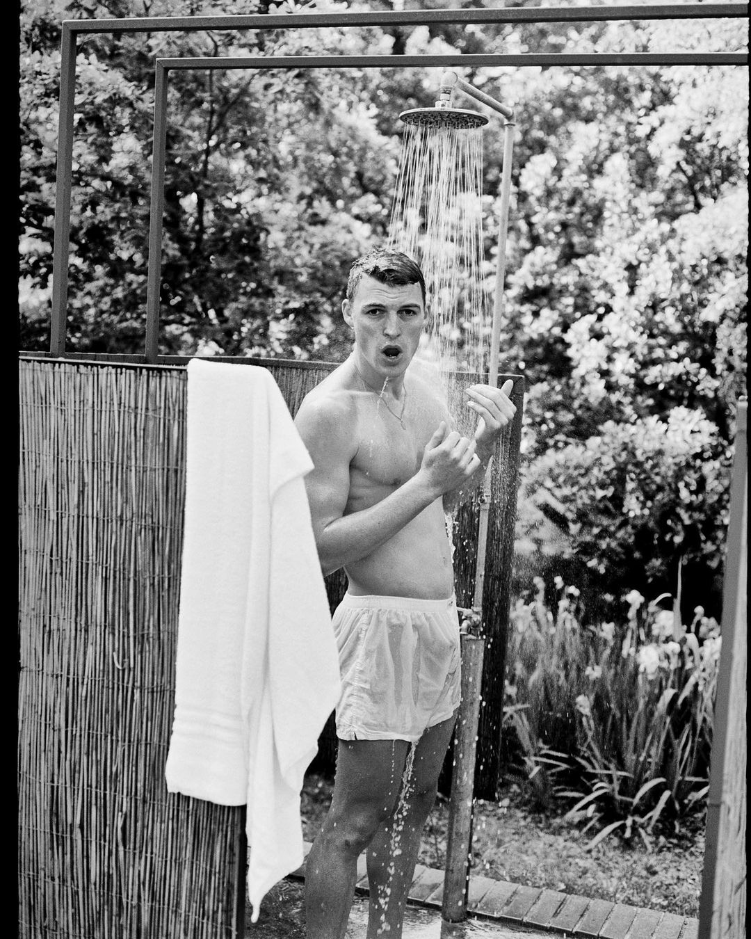 Monochrome outdoor shot of a male figure under a shower, with water cascading down, capturing a candid, refreshing moment near a fence.