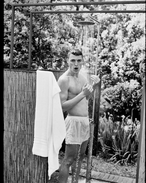Monochrome outdoor shot of a male figure under a shower, with water cascading down, capturing a candid, refreshing moment near a fence.