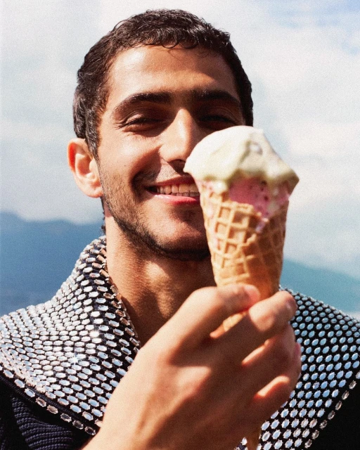 Smiling man in embellished jacket holds ice cream cone outdoors, portrait orientation, good lighting, clear focus, happy expression.