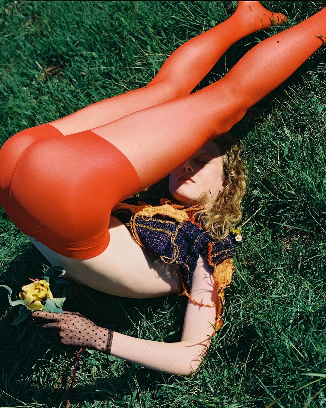 Young woman with orange tights laying in green grass holding a yellow rose, wearing a knit crop top and sheer dotted gloves.
