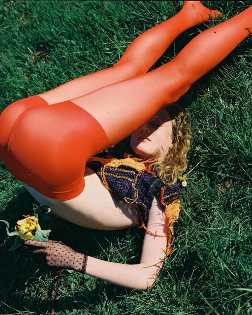 Young woman with orange tights laying in green grass holding a yellow rose, wearing a knit crop top and sheer dotted gloves.