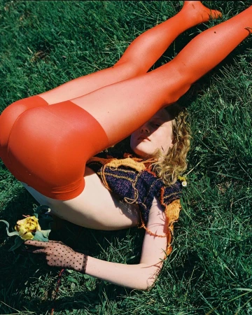 Young woman with orange tights laying in green grass holding a yellow rose, wearing a knit crop top and sheer dotted gloves.