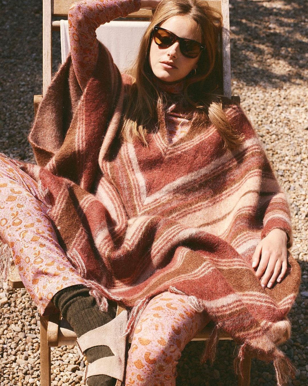 Adult woman in sunglasses and a poncho sits in the sun wearing printed pants and sandals in an outdoor setting with gravel ground.