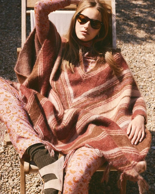 Adult woman in sunglasses and a poncho sits in the sun wearing printed pants and sandals in an outdoor setting with gravel ground.