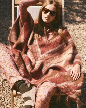 Adult woman in sunglasses and a poncho sits in the sun wearing printed pants and sandals in an outdoor setting with gravel ground.