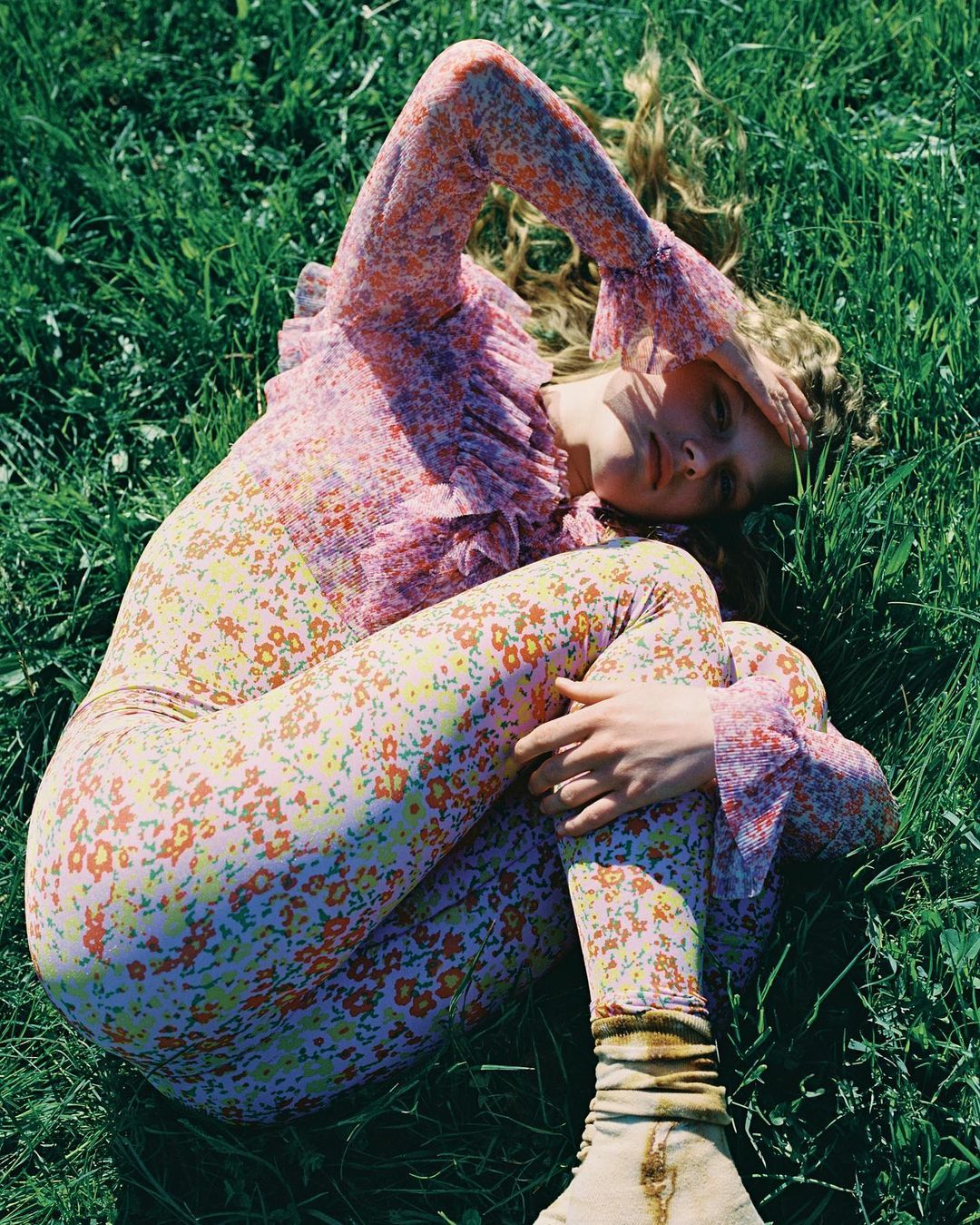 Woman in floral print top and leggings lies in green grass with hand over face in a close-up, eye-level shot; summer fashion editorial.