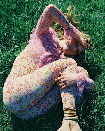 Woman in floral print top and leggings lies in green grass with hand over face in a close-up, eye-level shot; summer fashion editorial.