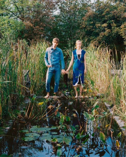 A couple stands barefoot in a natural pond, surrounded by tall reeds and greenery, wearing denim and a blue floral dress, holding hands.