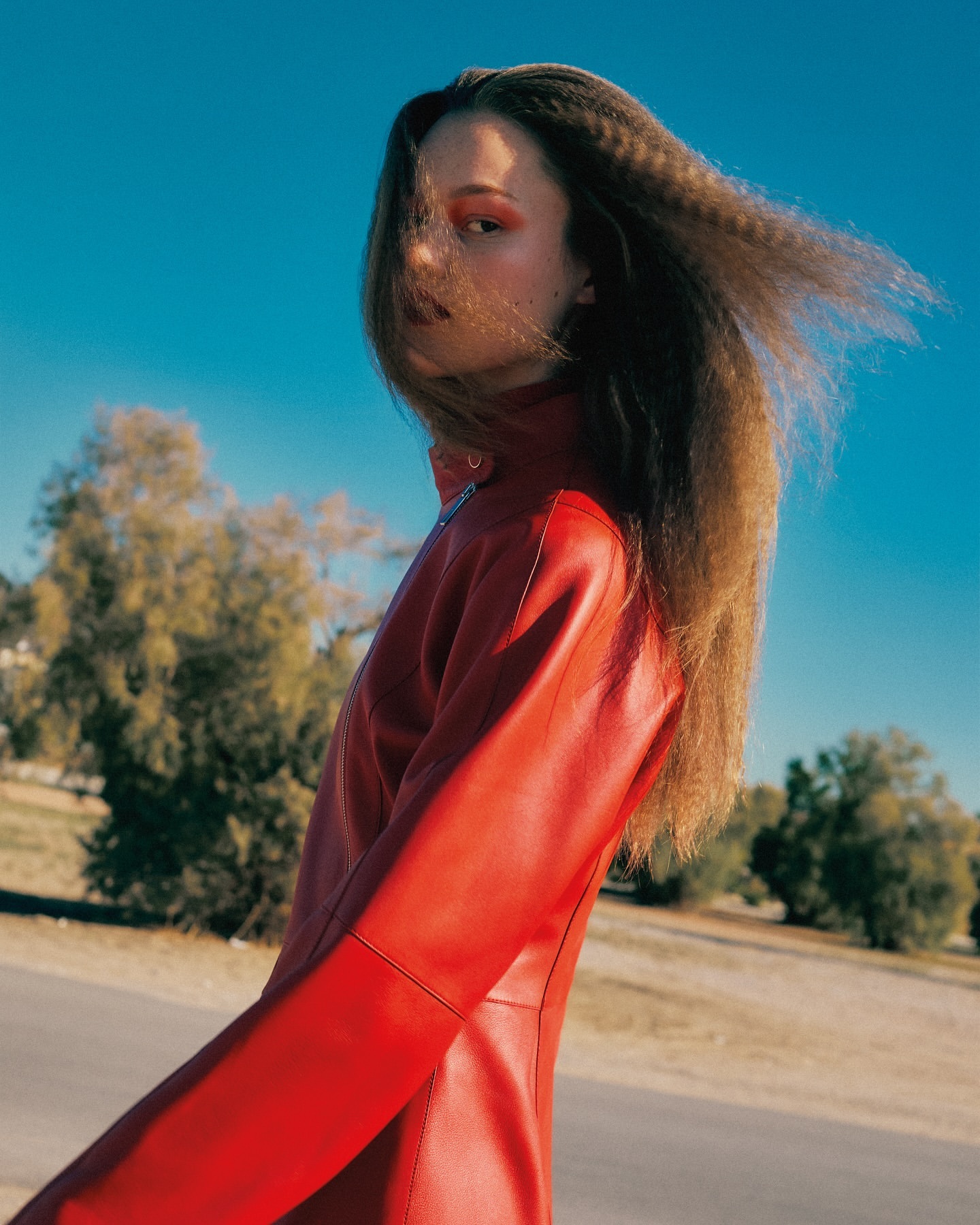 Woman in a red leather dress posing outdoors under a blue sky with trees in the background. Crimped long hair and make-up.