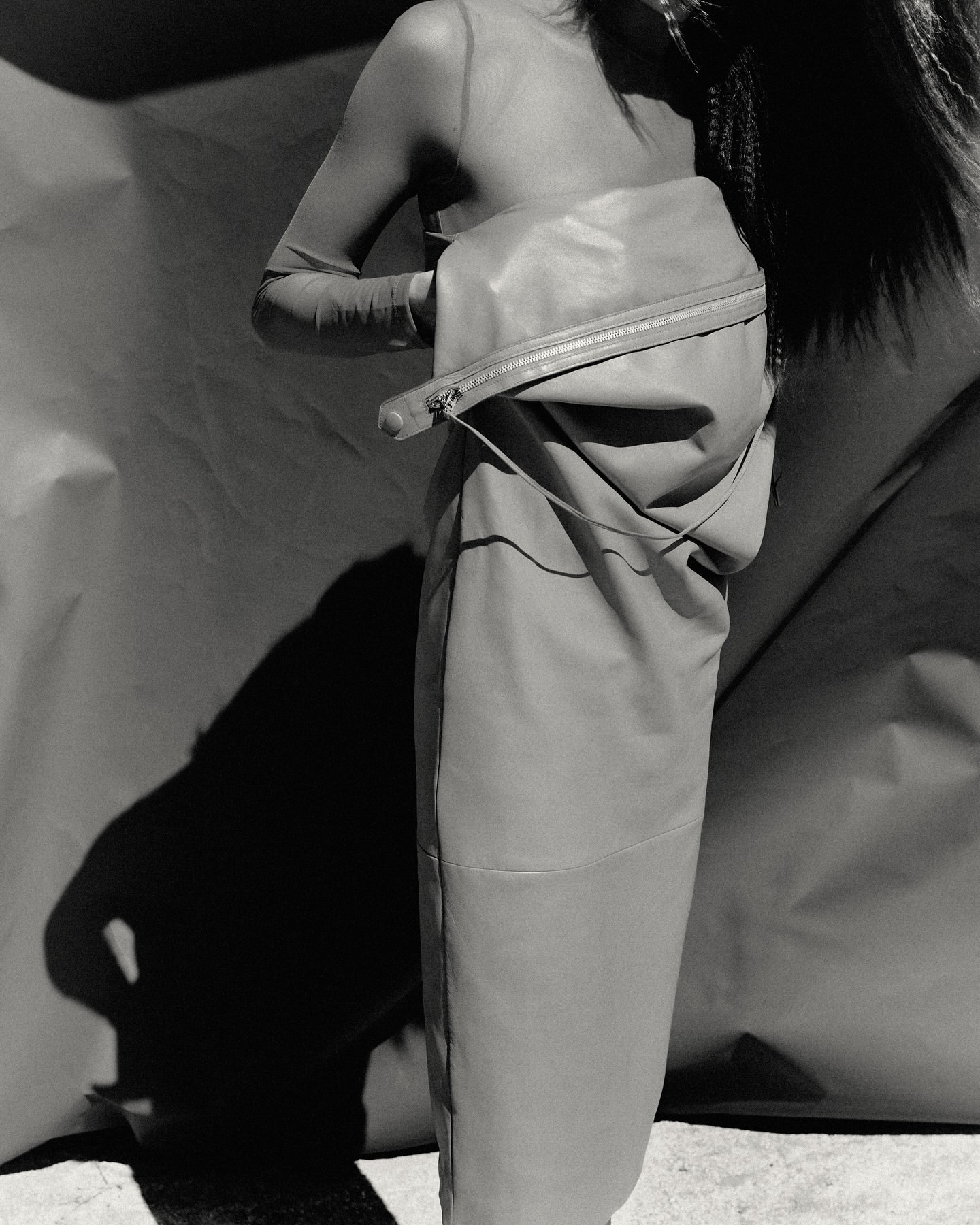 Monochrome fashion shot features an adult female posing with a large leather-like bag, styled with a translucent top, and dramatic shadow.