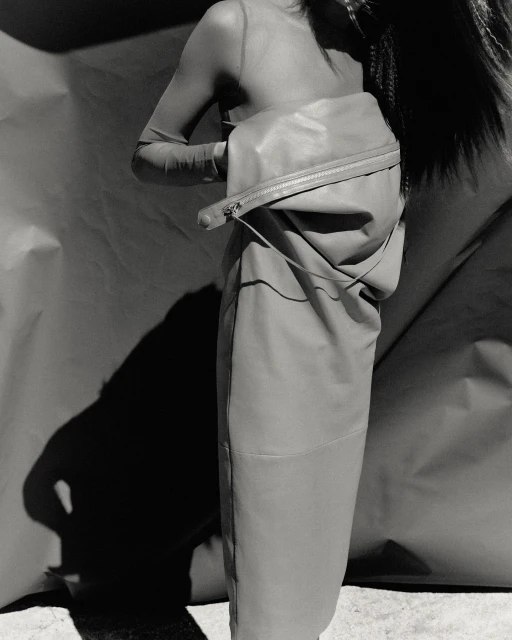 Monochrome fashion shot features an adult female posing with a large leather-like bag, styled with a translucent top, and dramatic shadow.