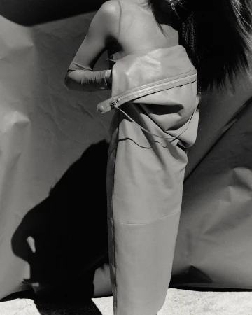 Monochrome fashion shot features an adult female posing with a large leather-like bag, styled with a translucent top, and dramatic shadow.