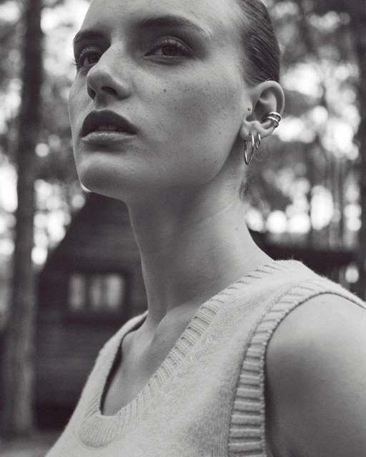 Monochromatic portrait of a young adult female with silver hoop earrings, wearing a sweater vest in an outdoor, wooded setting.