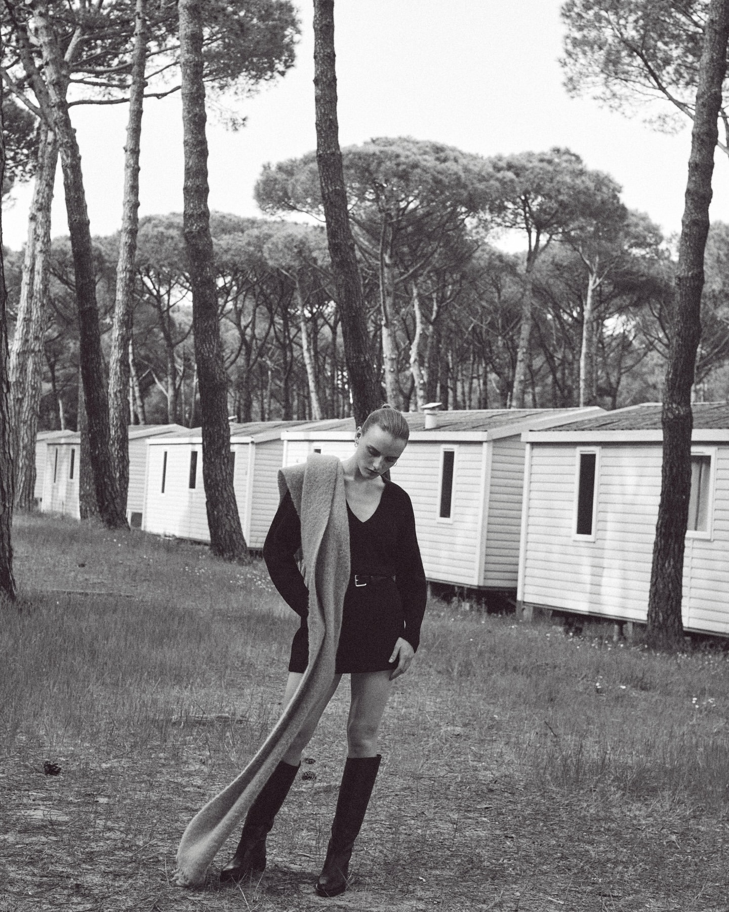 Monochrome fashion shot of a female model in a mini dress and boots, posing with a scarf in a wooded area with cabins.