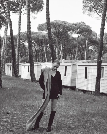 Monochrome fashion shot of a female model in a mini dress and boots, posing with a scarf in a wooded area with cabins.