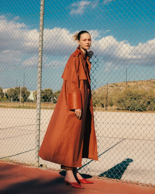 Fashion shot of an adult female wearing a long orange coat and red shoes standing in front of a tennis court and a chain-link fence.