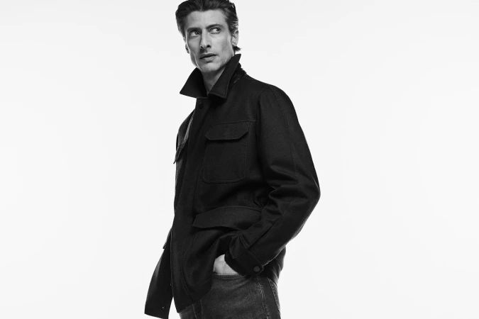 Man wearing a dark utility jacket and jeans looking over his shoulder against a plain white background