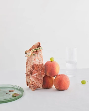 Still life photo featuring a snake-shaped bracelet on a marble block, peaches, grapes, and a glass plate against a white background.