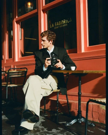 Man in a black blazer and light trousers seated at a cafe table holding a black camera in Milan by Alessandro Burzigotti
