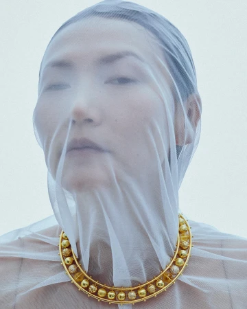 Close-up portrait of a woman wearing a sheer white veil over her face and a prominent gold beaded necklace.