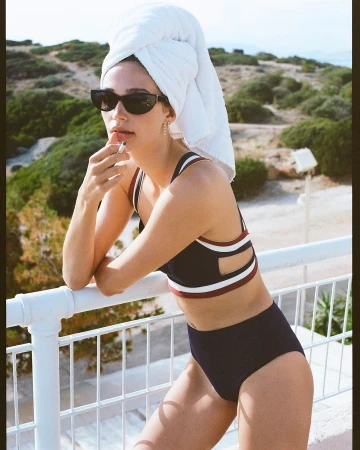 Woman in dark bikini and sunglasses with white bath towel on head leaning on a white railing outdoors.