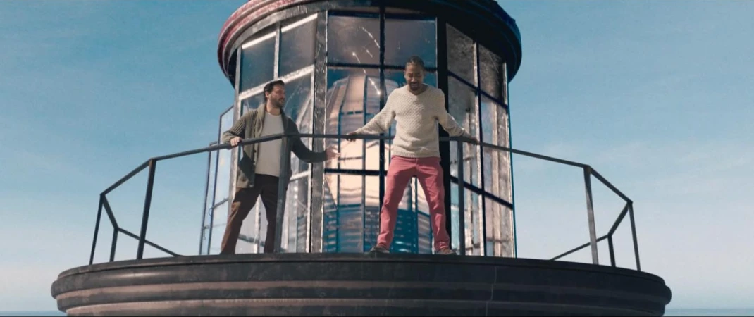Two adult men stand on a lighthouse observation deck on a sunny day, enjoying the view. The men are wearing casual attire.