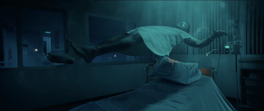 Adult male floats above a hospital bed, wearing patient gown in a dark hospital room with a window and medical equipment.
