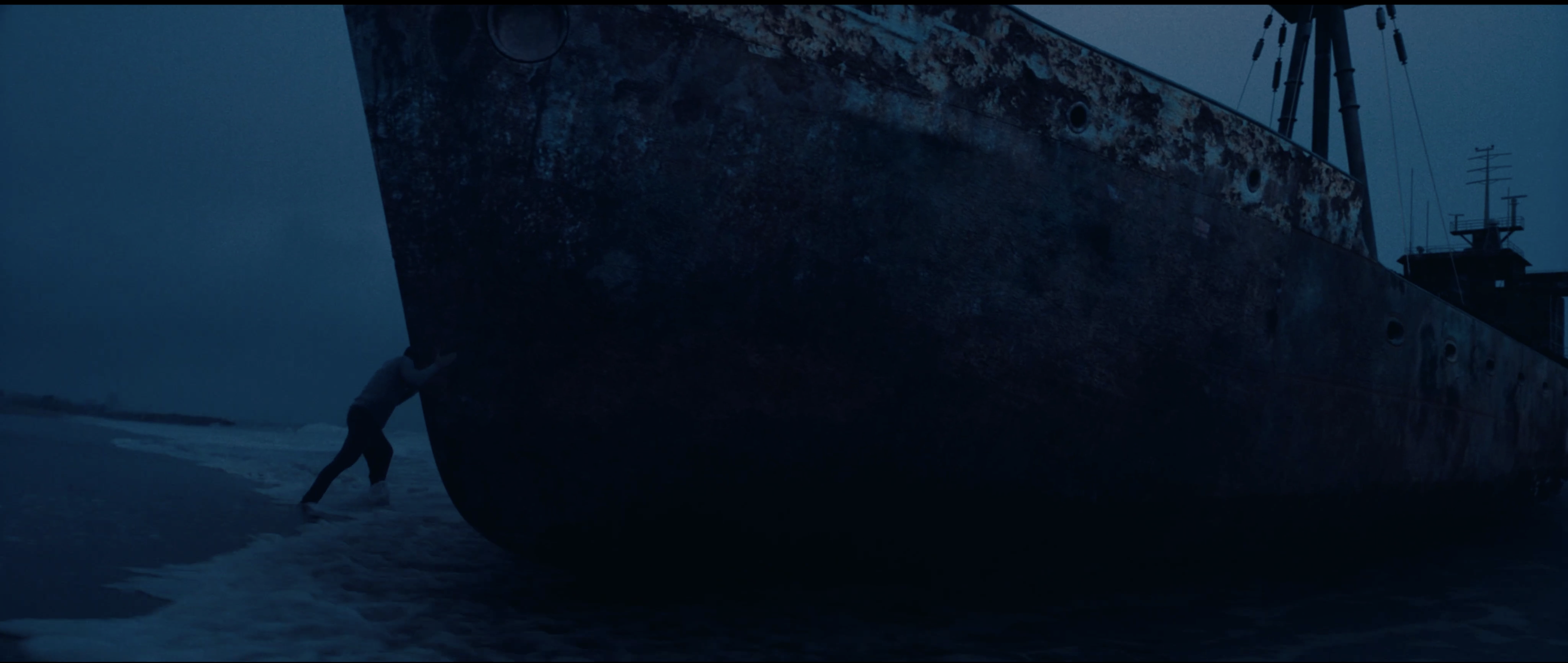 A lone figure pushes against the hull of a large, rusty shipwreck on a sandy beach at dusk, with a somber, moody atmosphere.