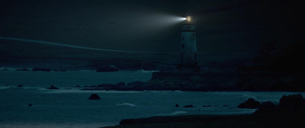 Lighthouse stands tall against the dark night sky on a rocky coast with ocean waves, illuminated by a bright light atop the tower.