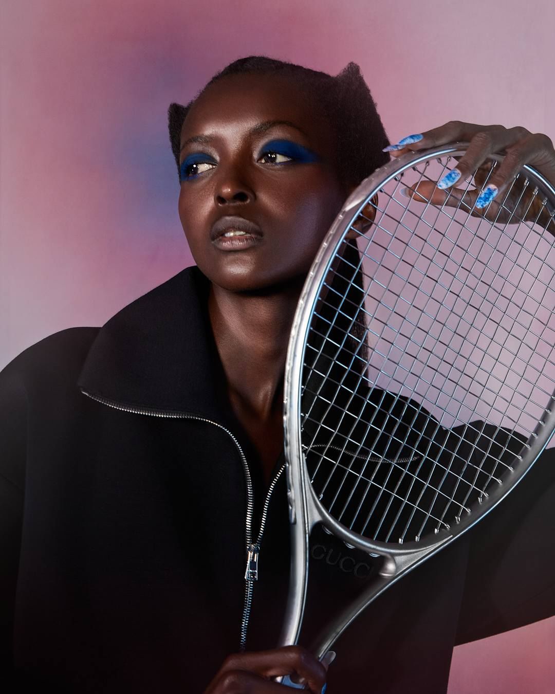 Dark-skinned woman poses indoors with tennis racket, wearing blue eyeshadow and a black jacket against a pink and blue backdrop.
